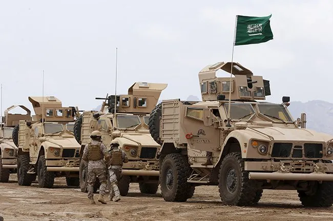 Saudi-led coalition armored vehicles and soldiers under a Saudi flag in the desert