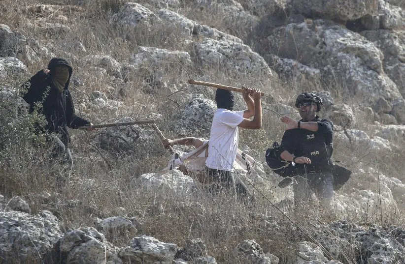 Masked settler wielding a stick attacks a person as a press-helmeted photographer looks on, West Bank hillside