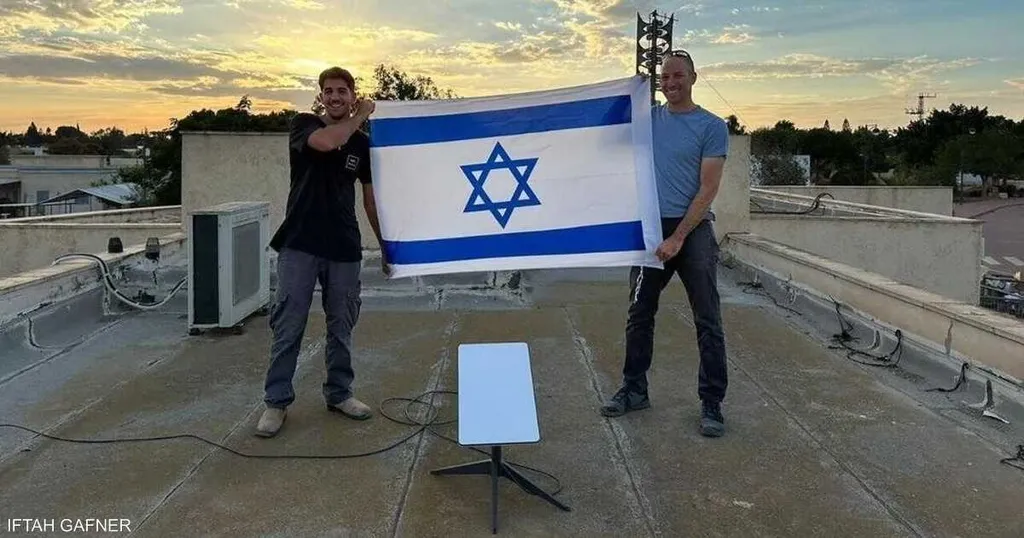 Two men hold an Israeli flag on a rooftop beside a Starlink terminal dish