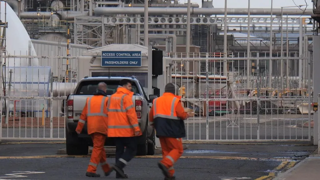 Workers in hi-vis jackets enter a refinery through an access control gate